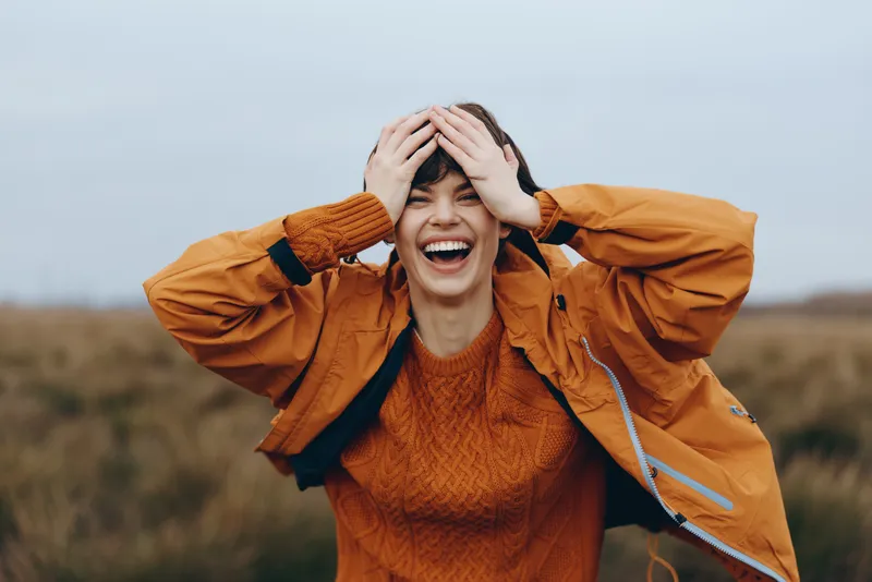 Woman lifestyle outdoors in a field, orange jacket and orange sweater, laughter as she covers her head with hands, joyful stress relief on a cloudy windy day