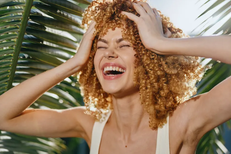 Laughing woman with curly hair enjoying tropical sun outdoors near palm leaves, expressing joy and carefree summer vibes in warm natural light.