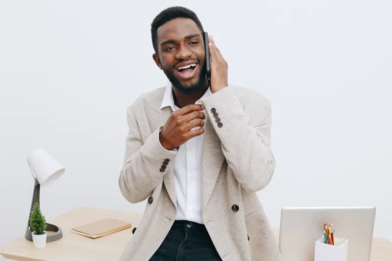 Confident smiling man with dark skin, beard, dressed in beige blazer and white shirt, talking on mobile phone, sitting at modern home office desk with laptop and stationery, indoor environment, casual