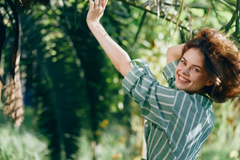 A cheerful woman outdoors wears a striped shirt, smiling as she reaches up to grip a rope, surrounded by lush green trees and sunlight, capturing a playful moment of freedom and nature