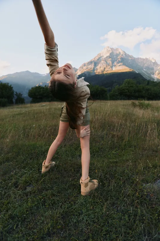 Woman performing a graceful backbend outdoors in a grassy field with mountains in the background, expressing freedom and playfulness on a sunny day under bright blue sky.