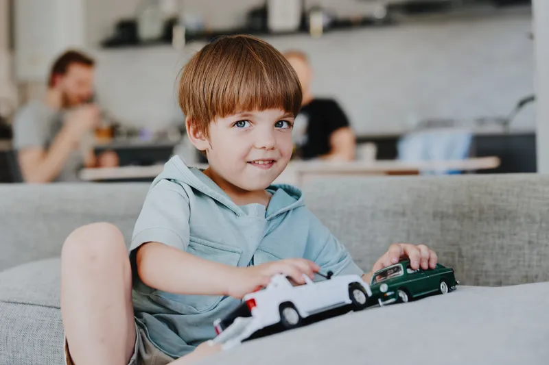 Smiling boy playing with toy cars, cheerful atmosphere, cozy home interior, family time, vibrant colors, adorable expression.