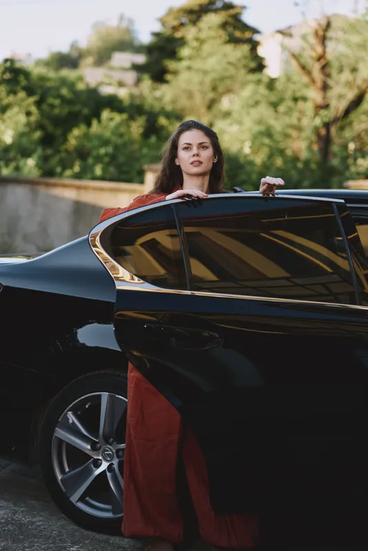 A stylish young woman in a red dress leans from a sleek black car, gazing at the camera with a calm, confident expression on a sunlit city street, suggesting luxury and independence