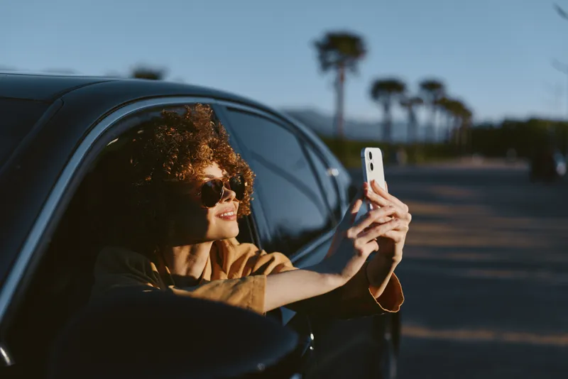 A woman in a car takes a selfie, enjoying a sunny drive, captured in retro film color tones. A candid lifestyle moment highlighting carefree travel and personal expression.