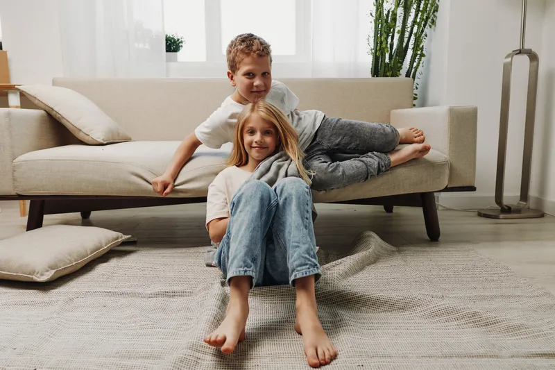 Two happy children relax in bright living room, barefoot on cozy carpet near modern sofa. Concept of family, childhood, home comfort and sibling bond.