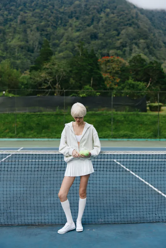 Young female athlete in white sportswear holding a green tennis ball on a sunny outdoor court surrounded by lush greenery, conveying a healthy lifestyle and active sports