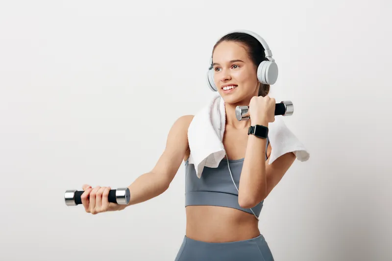 Woman fitness enthusiast in a gym, gray sports bra and leggings, lifting dumbbells with a towel around the neck and headphones, showcasing energetic workout.
