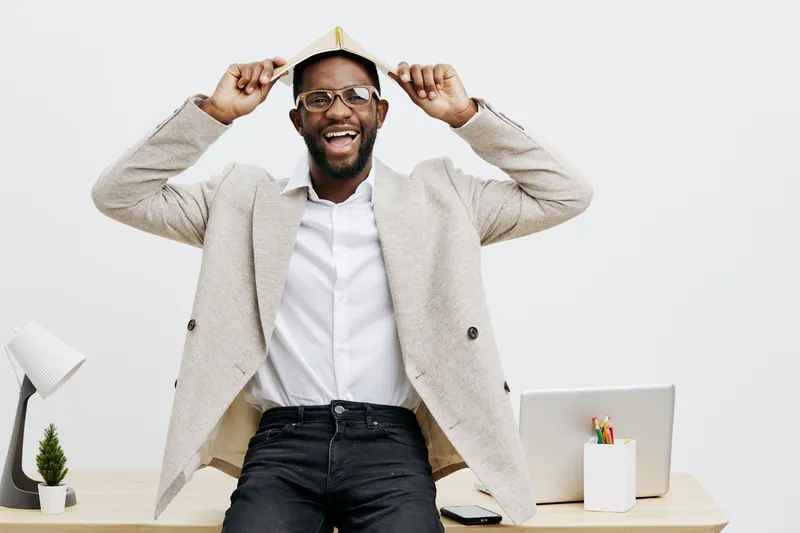 Joyful young African American man celebrating birthday or special occasion, holding a party hat on his head and smiling brightly, in casual attire, in an office or home workspace, light background. Pe