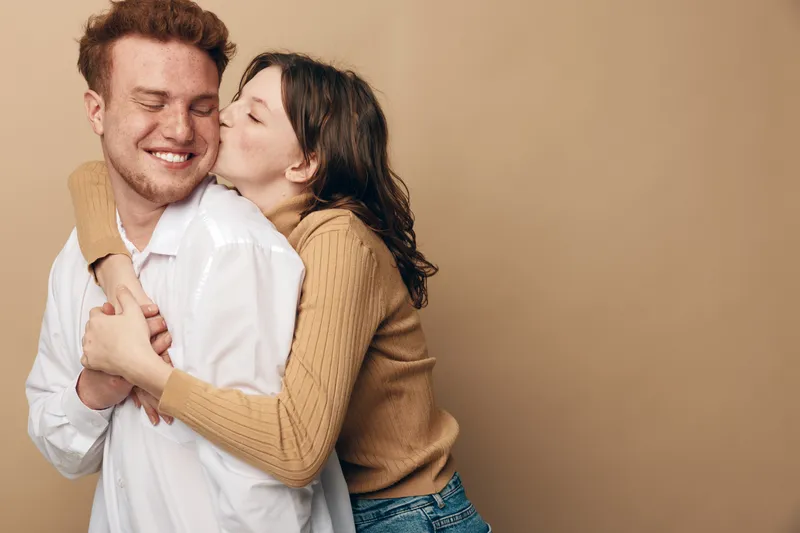 Happy young couple embraces and smiles in casual clothes on beige background. Woman kisses man on cheek showing affection and love. Concept of romance, relationship, and happiness.