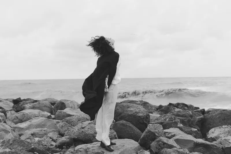 Couple embracing on a rocky sea shore, wind lifting coats and dress, black and white scene, intimate moment as waves crash against the rugged coastline, dramatic outdoor romance and moody atmosphere