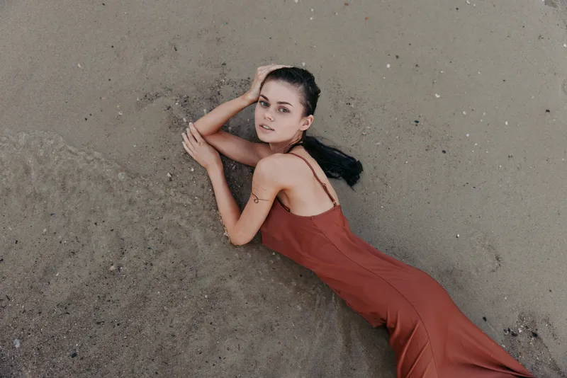A woman in a rust red dress lies on sandy ground, posing softly with her arms bent, creating a graceful, intimate portrait on a sunlit beach surface.