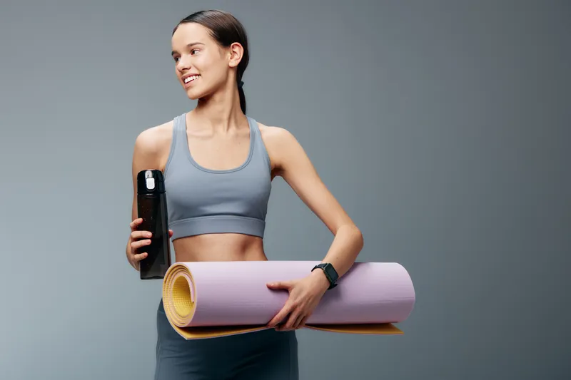 Fitness oriented woman in workout attire holds a water bottle and a rolled yoga mat, ready for training. Bright studio backdrop highlights strength, balance and motivation.