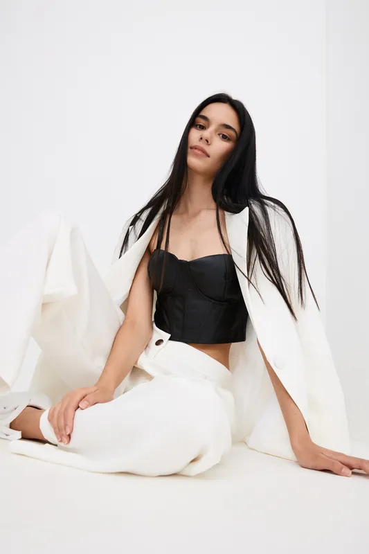 young woman in stylish white trousers and blazer sits relaxed against white background wearing a black corset top, showcasing modern minimalistic fashion and elegance