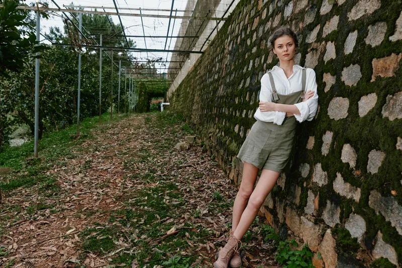 Young woman in olive green overalls and white shirt stands against a textured stone wall in a lush outdoor setting, exuding a relaxed and confident vibe