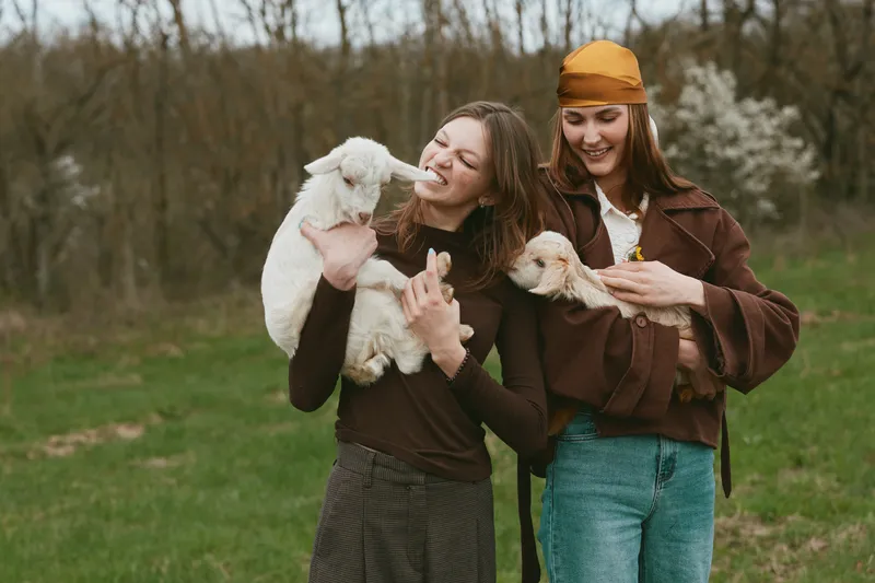 Two women outdoors in a park share a happy moment with goats and a kid, smiling, wearing casual outfits, a warm lifestyle scene highlighting friendship and animal joy.