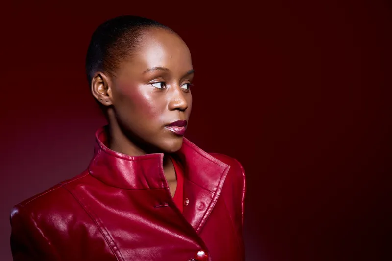 Portrait of a dark skinned African woman wearing a red leather jacket looking sideways with a serious expression. Studio shot with rich red background, fashion and beauty concept.