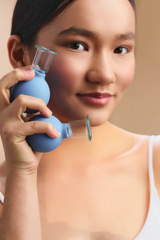 Asian woman is seen using a handheld facial suction device in a bright, clean space, smiling softly as she focuses on a soothing skincare routine and personal wellbeing.