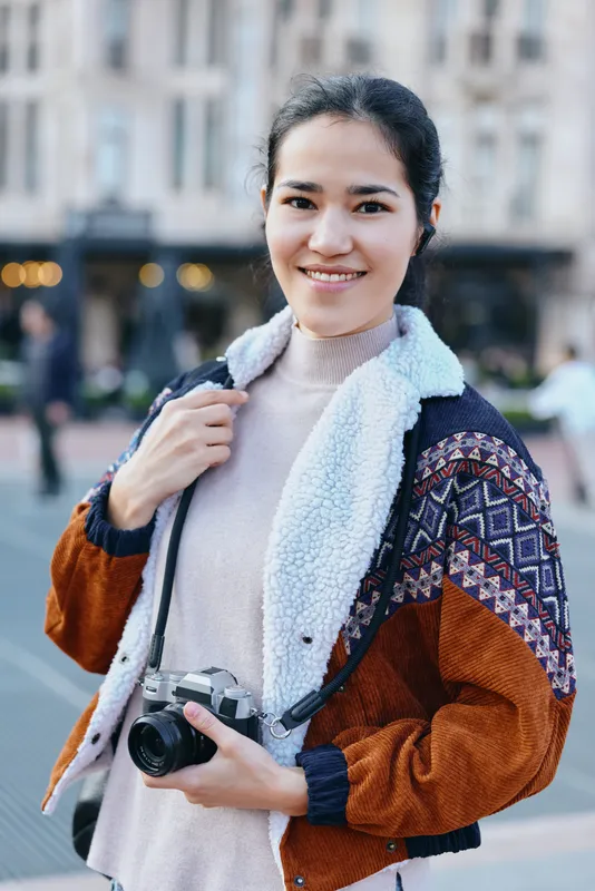 Smiling photographer standing on city street with camera in hand, casual jacket and soft sweater, urban backdrop, candid style, everyday creativity, travel and street photography vibe