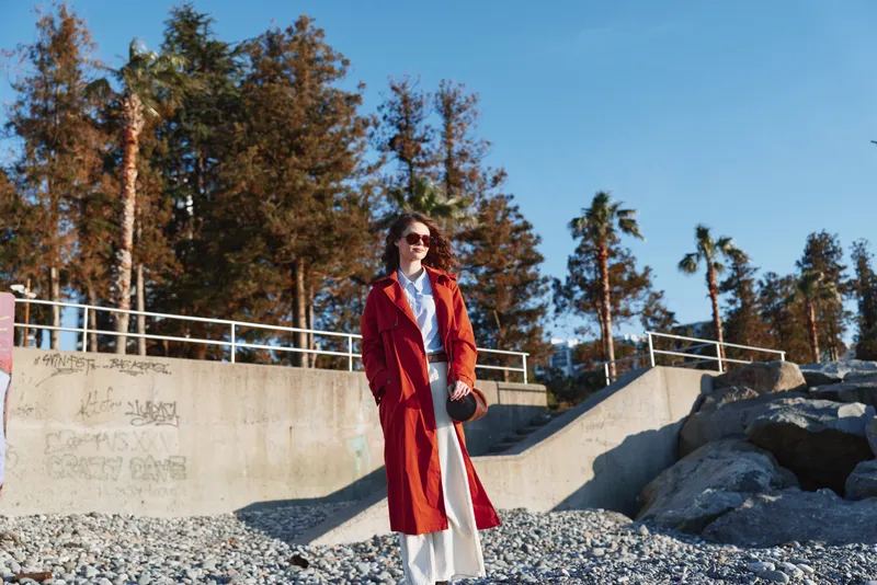 A woman in a long red coat and white pants stands on a rocky beach near concrete stairs with palm trees and clear sky in the background on a sunny day.