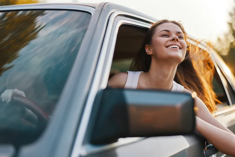 Happy young woman enjoying a road trip with her head out of a car window, symbolizing freedom and adventure during a sunny day outdoors.
