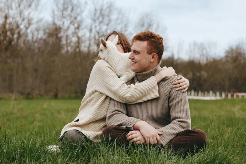 Smiling couple sitting on grass outdoors, woman embracing man while a curious goat kid nudges them. Cozy sweaters, soft daylight, open field, warm connection, simple lifestyle moment, natural