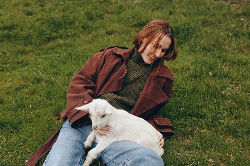 Lifestyle scene of a young person on grass with a white goat kid, sharing a tender moment, relaxed clothing, natural light, peaceful meadow mood, simple outdoor companionship and calm joy