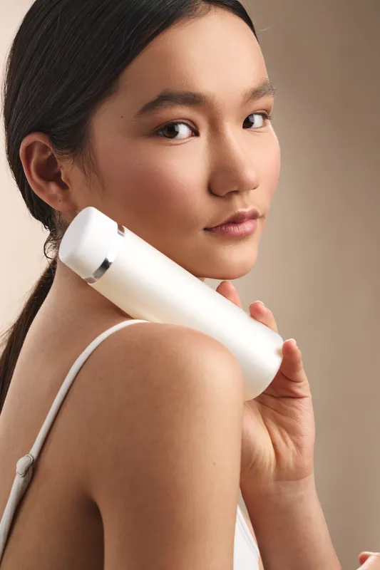 asian woman holds a sleek skincare device against her shoulder, wearing a light top, and gazes at the camera with calm confidence, conveying a modern, minimal beauty routine in a clean studio setting.