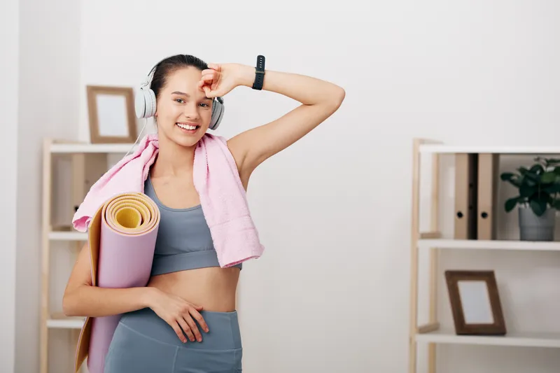 Smiling woman in athletic wear with headphones, carrying a yoga mat and towel after a workout in a bright home studio, ready for cooldown and stretch.