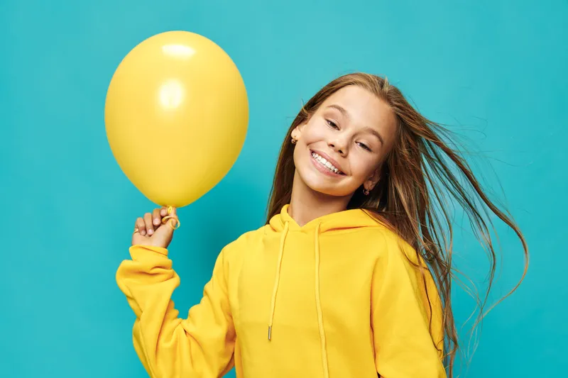 Girl smiling and holding yellow balloon with yellow hoodie on blue background. Portrait of happy young female with long hair in casual clothing for celebration or party.