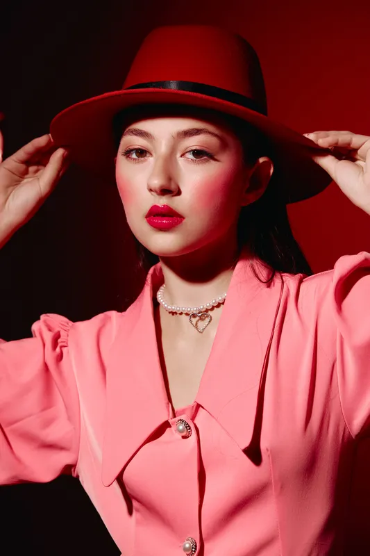 Young woman in red hat and pink jacket poses with hands near head. Studio portrait focusing on dramatic makeup and bold colors. Concept of beauty creative fashion and elegance.