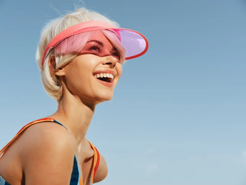 Joyful portrait of a smiling woman in a pink visor enjoying a clear sunny day, capturing vibrant energy and carefree summer vibe under a bright blue sky.