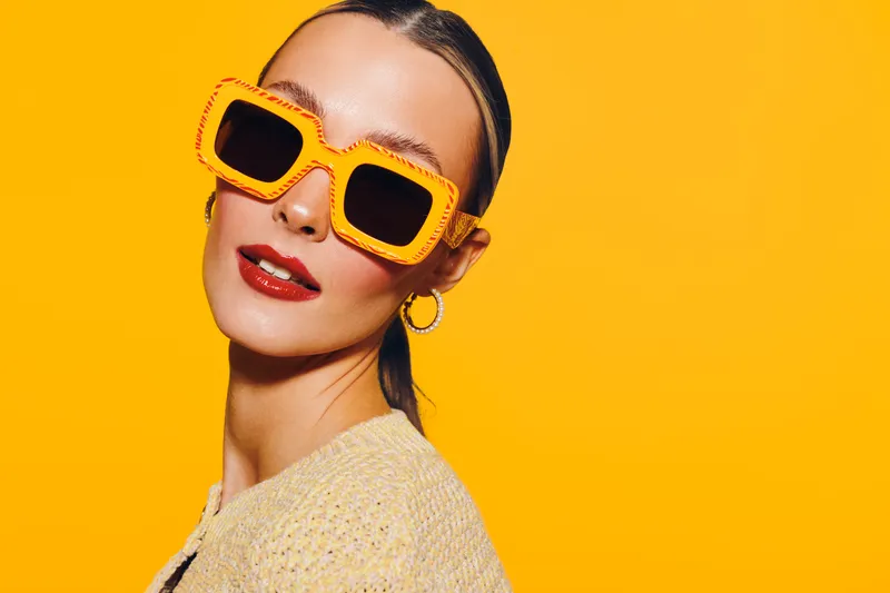 Stylish woman with slicked hair wearing large orange sunglasses and red lipstick poses against bright yellow background. Fashion concept with bold colors and confident expression.
