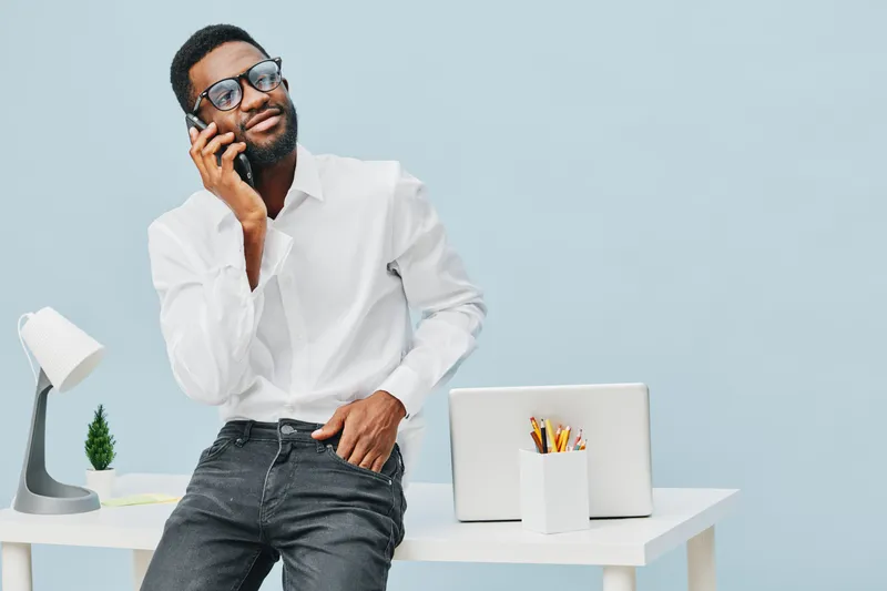 Full body portrait of a young stylish man with glasses talking on the mobile phone while standing near a desk with laptop and stationery, casual clothing, modern office interior, light background, lif