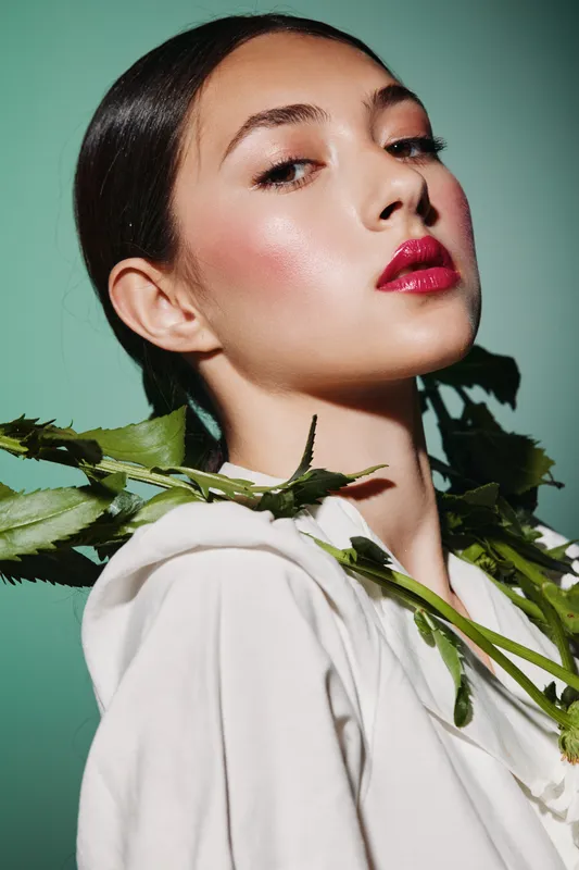 Close-up portrait of a fashion model with glossy makeup and bright lips posing with green leaves on her shoulder against a soft teal background, creative beauty concept.