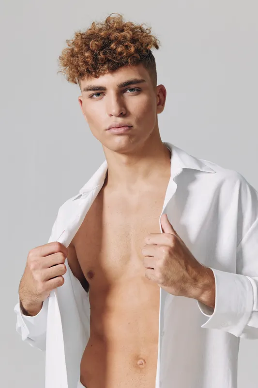 Young man with curly hair posing shirtless wearing an open white shirt against a plain background. Confident male model with natural skin and casual style in studio.