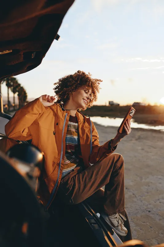 Woman lifestyle travel on beach at sunset, sitting by a car, checking smartphone and phone in a casual jacket. Curly hair, warm sunlight, calm leisure.