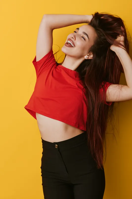 Young woman with long hair wearing red crop top and black pants posing happily with hands in hair against yellow background. Smiling, joyful and carefree female model in casual outfit.