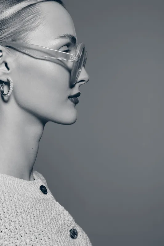 Profile of a young white woman with fair skin wearing stylish transparent sunglasses and earrings on a neutral background. Fashion and beauty concept.