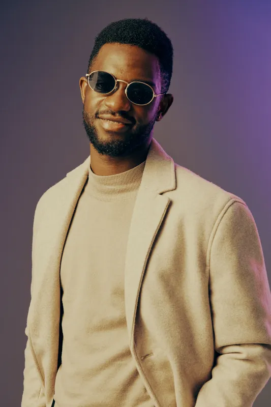 Stylish man in sunglasses wearing a beige coat with a smile, set against a colorful gradient background, representing modern fashion and confidence