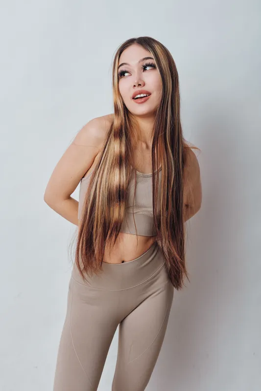 Young woman with long straight hair wearing beige sportswear poses and looks sideways on white background, fitness lifestyle, casual style, confidence and youth concept.