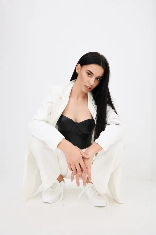 Woman in white suit and black corset squatting on white background with relaxed posture, long dark hair, and casual white sneakers, modern fashion style.