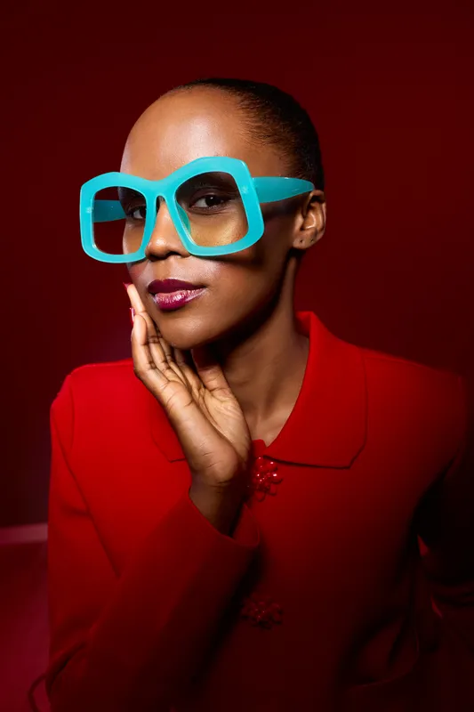 Stylish African woman with dark skin wears large blue glasses and red outfit. She poses thoughtfully with hand near face against deep red background. Concept of fashion and elegance.
