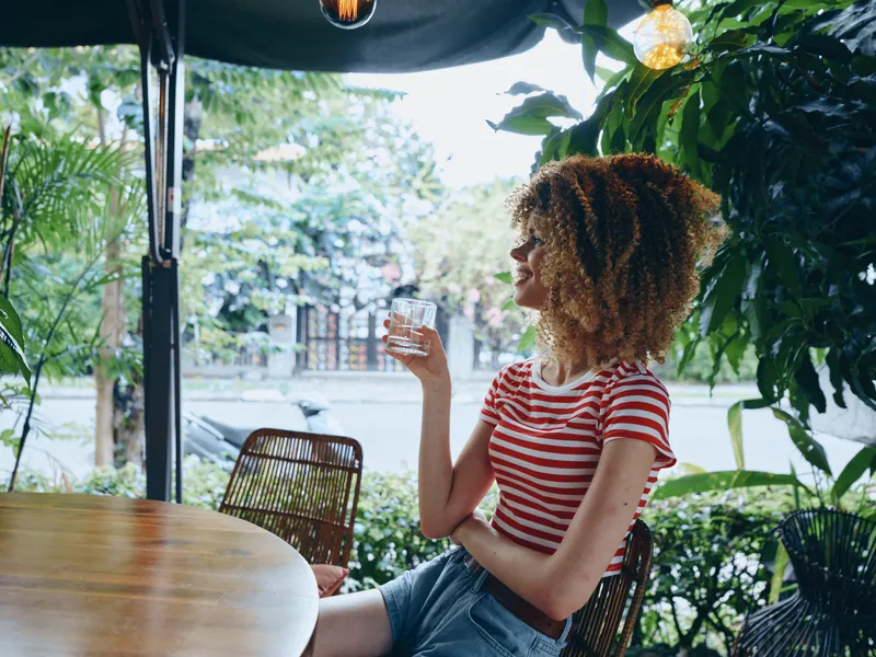 A woman with curly hair sits at an outdoor cafe, wearing a striped tee, holding a glass and enjoying a refreshing moment in warm natural light, with green plants surrounding the scene.