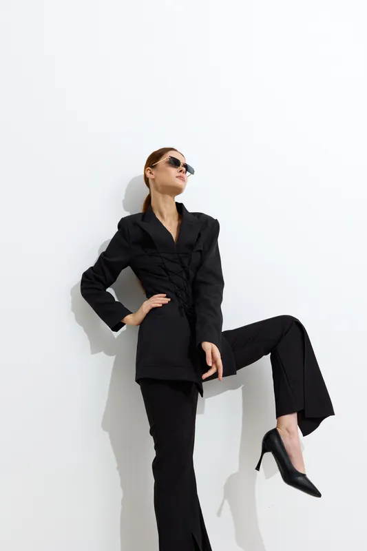 Stylish woman in black suit and sunglasses posing confidently against a white background with dramatic shadows, showcasing modern fashion and strength.