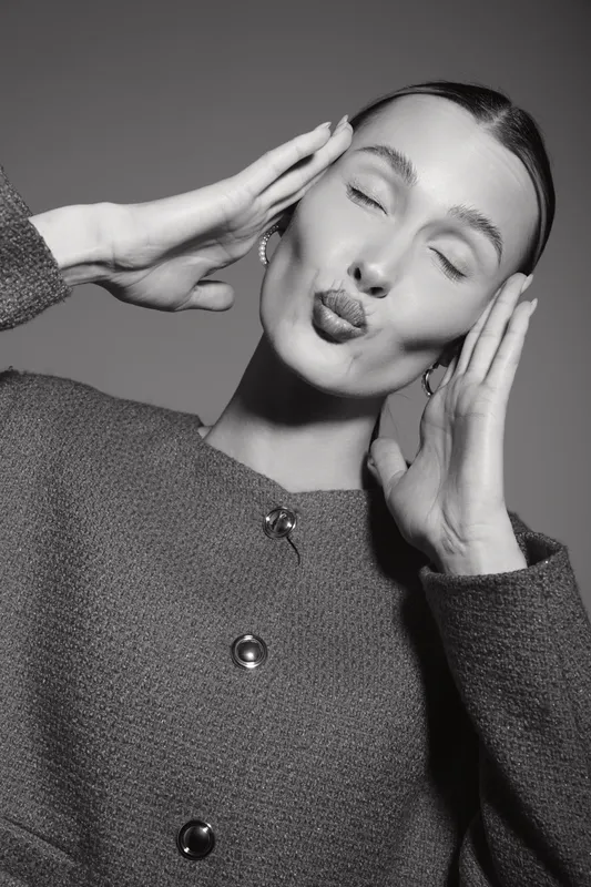 White woman with light skin and dark hair makes playful face with puckered lips and closed eyes wearing textured jacket in studio. Concept of fun, fashion, and portrait.