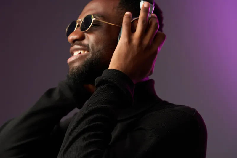 Smiling Black man enjoying music with headphones, wearing sunglasses, in a stylish black outfit with a colorful background