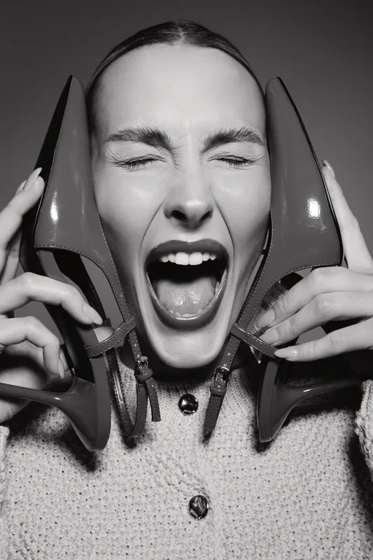 Young white woman with light skin holds high heel shoes near face and screams with closed eyes in studio.