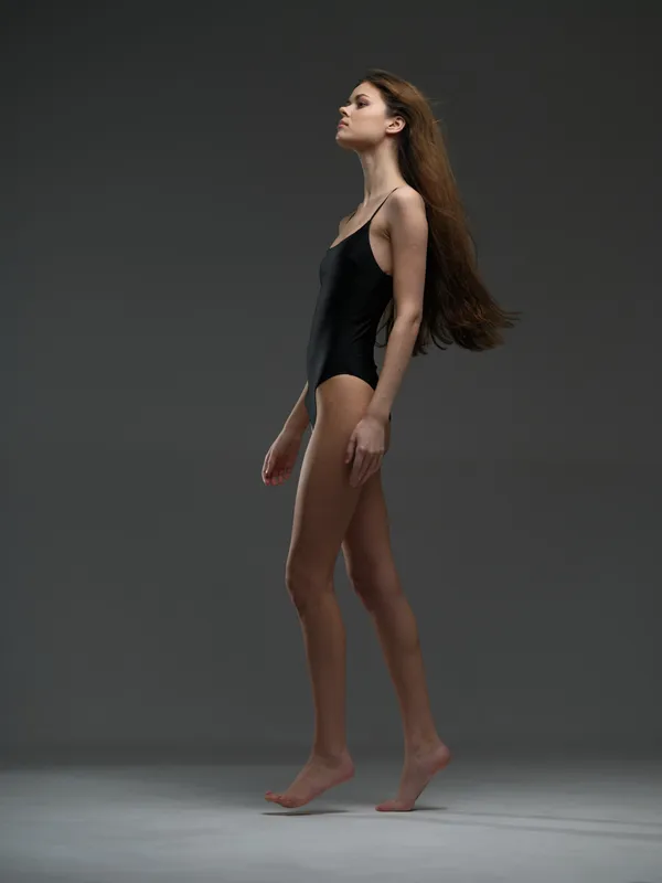 Standing woman in black swimsuit with long hair on gray background, elegant pose, minimalistic studio setting