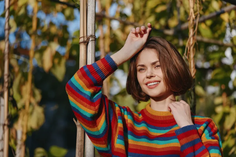 Woman in a striped sweater smiling and leaning on a wooden trellis in a sunlit garden, short bob hairstyle, casual outfit, relaxed cheerful portrait for lifestyle and fashion.