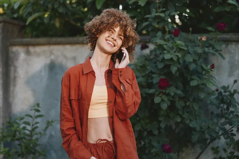 Casual woman with curly hair wearing a red jacket and crop top, talking on a phone in a garden. Bright natural light creates a relaxed outdoor lifestyle moment with warm, intimate mood